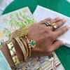 A close-up of a hand wearing multiple gold-toned bracelets and rings, resting on a patterned magazine page over a green surface.