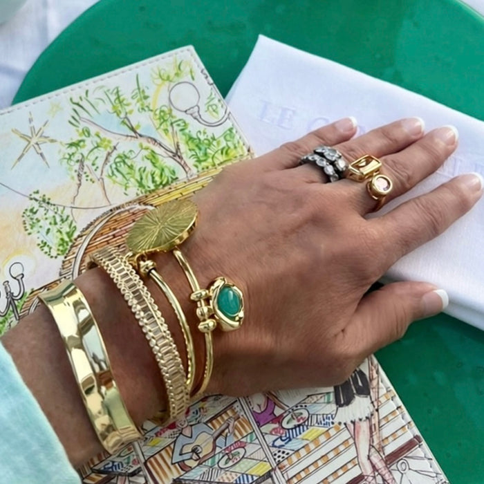 A close-up of a hand wearing multiple gold-toned bracelets and rings, resting on a patterned magazine page over a green surface.