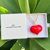 Red heart-shaped pendant on a gold chain in a white box with 'The Christy F Collection' branding, surrounded by green leaves.