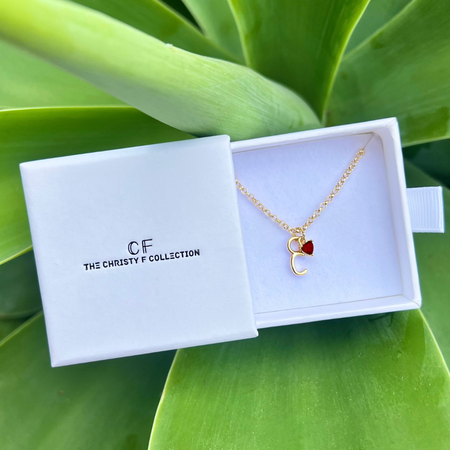 Gold necklace with a red heart pendant in a white box labeled 'CF The Christy F Collection' on green leaves.