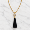 A gold chain necklace with a round bezel-set clear crystal pendant and a long black tassel drop, displayed on a white marble background.