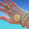 A close-up of a hand wearing multiple gold-toned jewelry pieces, including a large textured ring, a small pink-stoned ring, a round sunburst bracelet, and twisted bangles, set against a bright blue sky and turquoise ocean background.