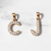Three gold initial pendant charms spelling out 'CJM' are arranged on a white marble surface. Each letter is paved with small clear stones.