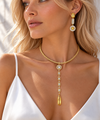 A close-up lifestyle shot of a blonde woman wearing a gold necklace with a teardrop pendant and matching gold drop earrings with clear stones.