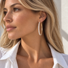 A close-up side profile of a woman with blonde hair wearing a large gold hoop earring set with clear pavé stones, paired with a white collared shirt.