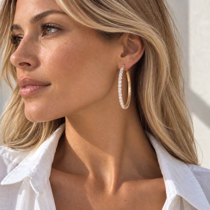 A close-up side profile of a woman with blonde hair wearing a large gold hoop earring set with clear pavé stones, paired with a white collared shirt.