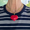 Red heart-shaped pendant on a gold chain necklace worn by a person in a striped shirt.
