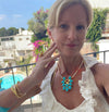 A woman wearing a turquoise and gold flower-shaped pendant necklace with a pearl chain, posing on a sunny balcony with a swimming pool and white buildings in the background.
