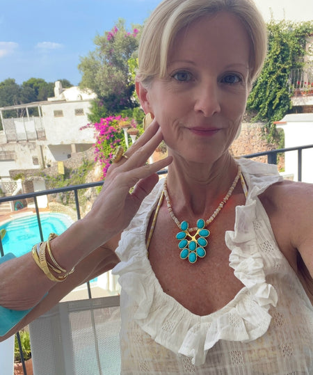 A woman wearing a turquoise and gold flower-shaped pendant necklace with a pearl chain, posing on a sunny balcony with a swimming pool and white buildings in the background.