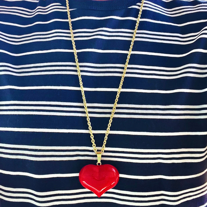 Red heart-shaped pendant on a gold chain against a blue and white striped shirt