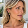 A close-up of a woman with blonde hair wearing a pair of drop earrings. The earrings feature a bright yellow top stone and a smaller pink bottom stone, both set in gold-toned metal.