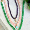 Three beaded necklaces in green, pink, and purple on a textured surface.