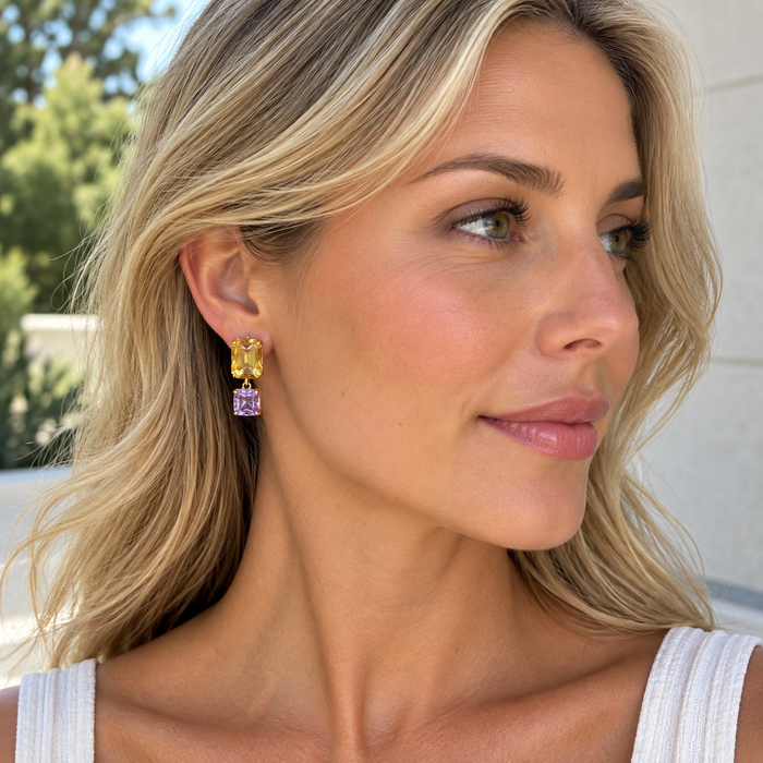 A close-up of a woman with blonde hair wearing a dangling earring with a yellow butterfly-shaped top and a purple rectangular drop.