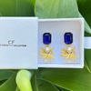 Gold earrings with blue gemstones in a white box labeled 'CF The Christy F Collection' on green leaves.