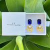 Gold earrings with blue gemstones in a white box labeled 'CF The Christy F Collection' on green leaves.