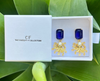Gold earrings with blue gemstones in a white box labeled 'CF The Christy F Collection' on green leaves.
