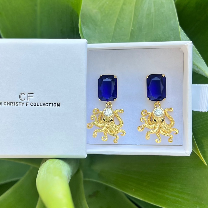 Gold earrings with blue gemstones in a white box labeled 'CF The Christy F Collection' on green leaves.