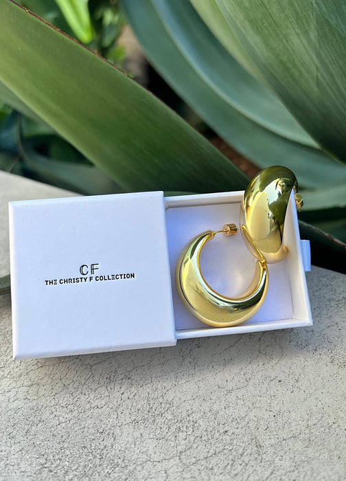 Gold hoop earrings in a white box with 'CF' branding, set against a natural background.