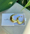 Gold hoop earrings in a white box with 'CF' branding, on a light surface with green leaves.