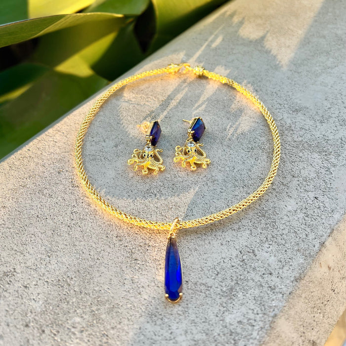 Gold necklace with blue teardrop pendant and matching earrings on a stone surface with green leaves in the background.