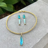 Gold necklace with blue pendant and matching earrings on a stone surface with green leaves in the background