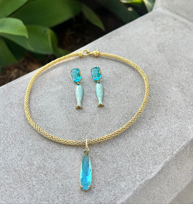 Gold necklace with blue pendant and matching earrings on a stone surface with green leaves in the background