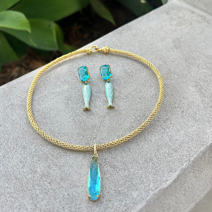 Gold necklace with blue pendant and matching earrings on a stone surface with green leaves in the background
