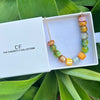Colorful beaded necklace in a white box with 'The Christy F Collection' branding, surrounded by green leaves.