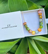 Colorful beaded necklace in a white box with 'The Christy F Collection' branding, surrounded by green leaves.