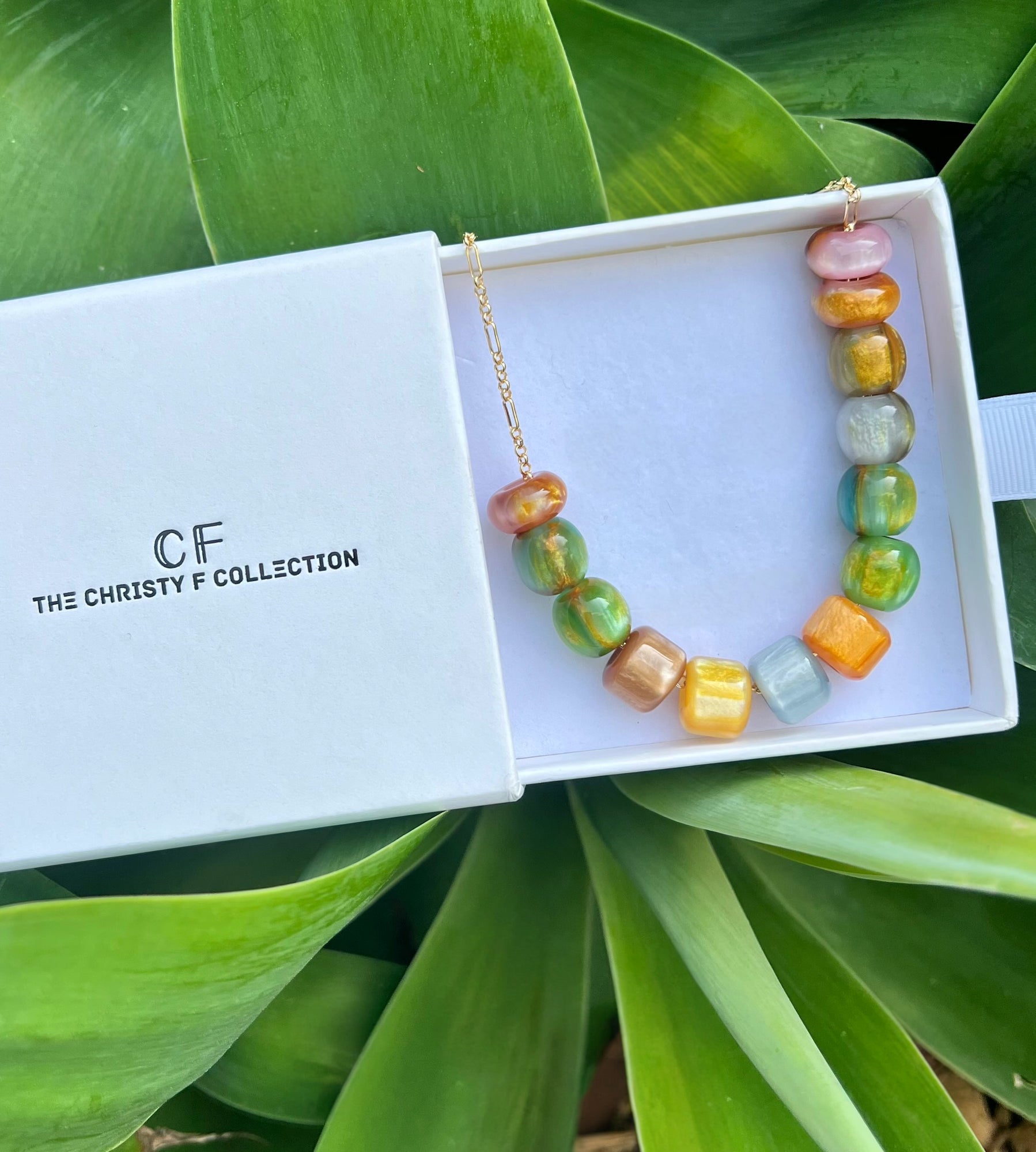 Colorful beaded necklace in a white box with 'The Christy F Collection' branding, surrounded by green leaves.