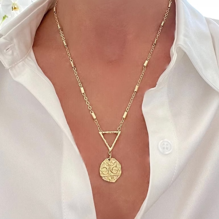 A gold-filled dainty tube chain necklace with a triangle connector and a hammered pendant, worn by a person with a white shirt.