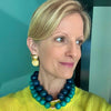 Woman wearing a blue and green beaded necklace and gold earrings against a teal wall.