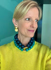 Woman wearing a blue and green beaded necklace and gold earrings against a teal wall.