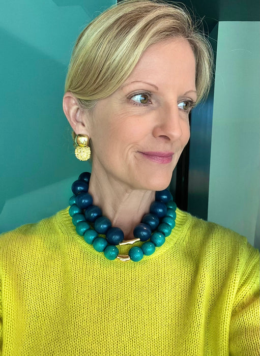 Woman wearing a blue and green beaded necklace and gold earrings against a teal wall.