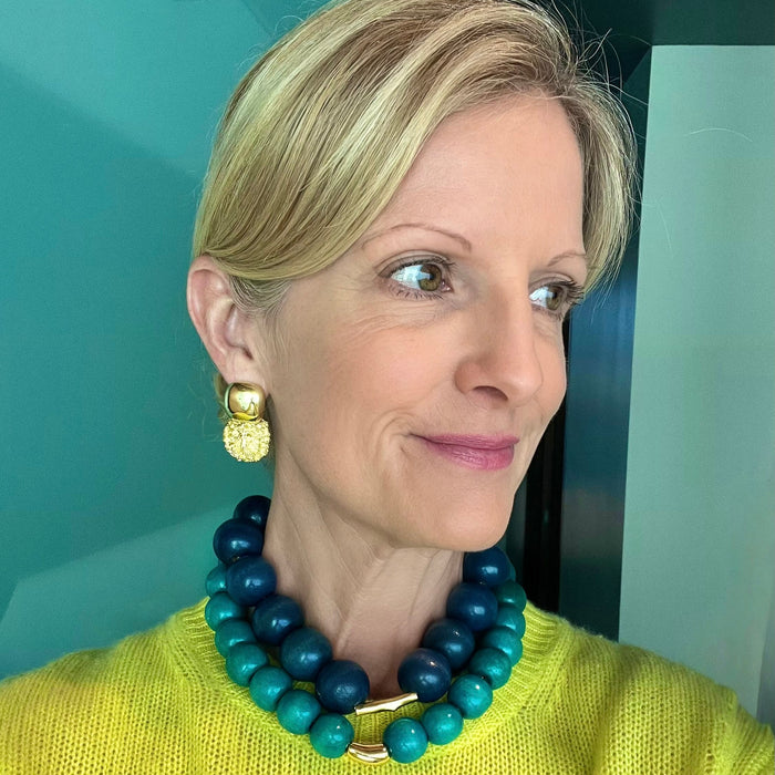 Woman wearing a blue and green beaded necklace and gold earrings against a teal wall.