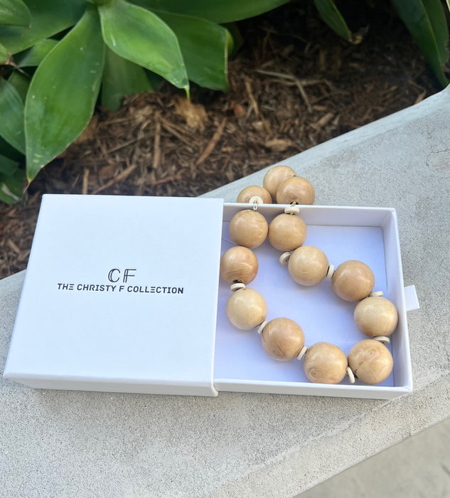 Wooden beaded necklace in a white box with 'The Christy F Collection' branding, placed on a stone surface with greenery in the background.