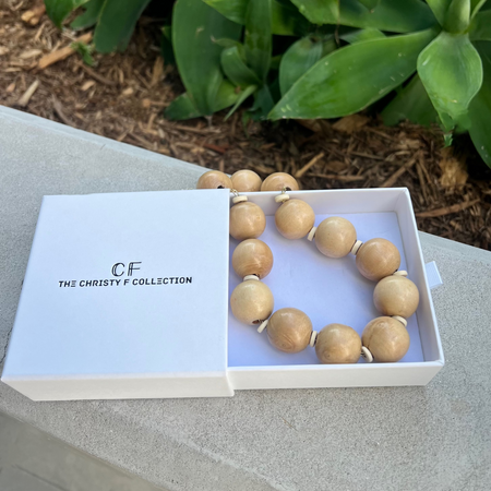 Wooden beaded necklace in a white box with 'GF The Christie F Collection' branding, placed on a stone surface with green foliage in the background.