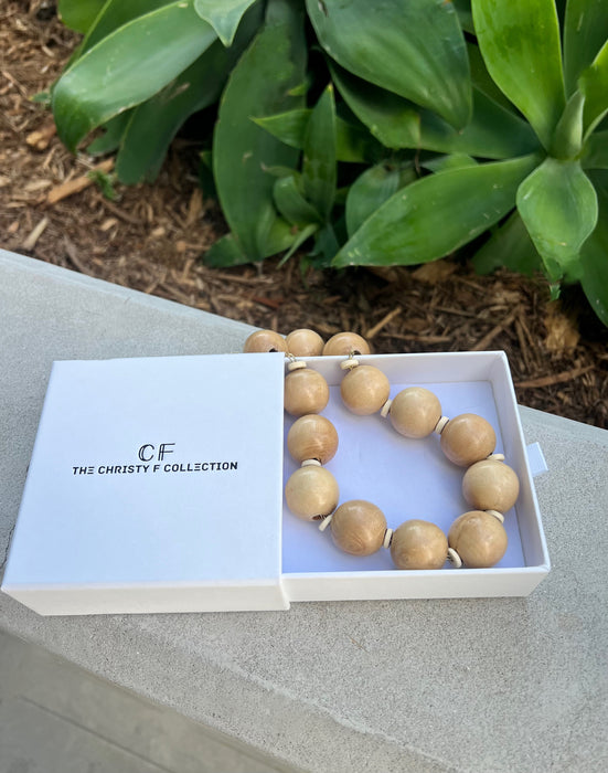 Wooden beaded necklace in a white box with 'GF The Christie F Collection' branding, placed on a stone surface with green foliage in the background.