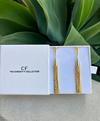 Gold tassel earrings in a white box with 'CF The Christy F Collection' branding, against a green leafy background.