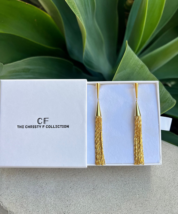 Gold tassel earrings in a white box with 'CF The Christy F Collection' branding, against a green leafy background.