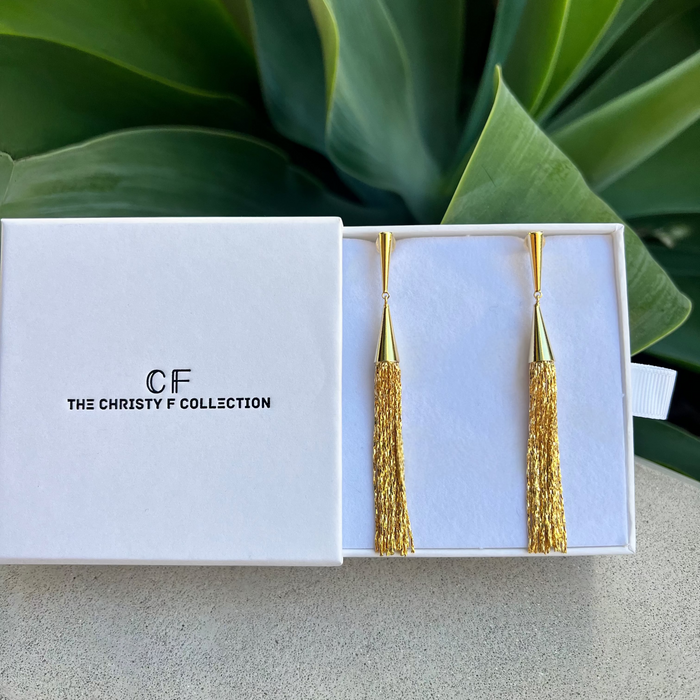 Gold tassel earrings in a white box with 'CF The Christy F Collection' branding, against a green leafy background.