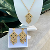 Gold jewelry set with necklace and earrings on a white stand and box against a green plant background.