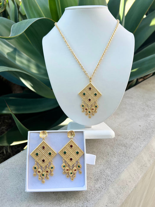 Gold jewelry set with necklace and earrings on a white stand and box against a green plant background.