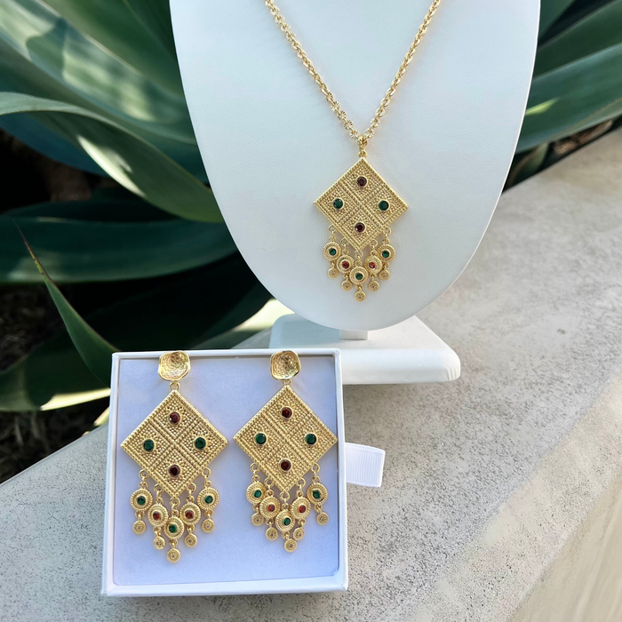 Gold jewelry set with necklace and earrings on a white stand and box against a green plant background.