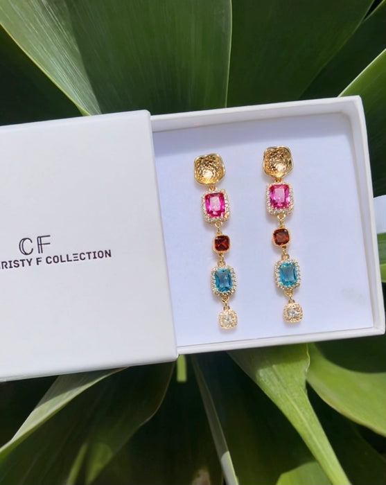 Colorful earrings in a white box with 'CF' branding, set against a green leafy background.