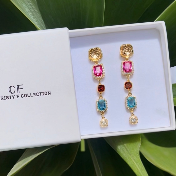 Colorful earrings in a white box with 'CF' branding, set against a green leafy background.