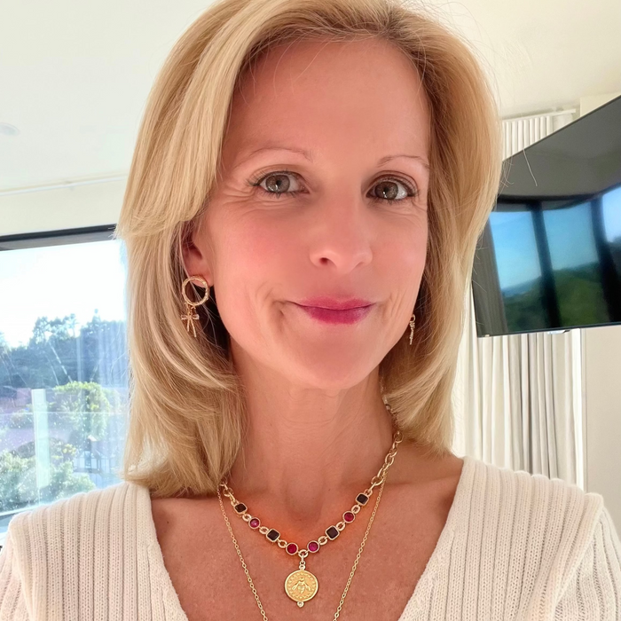 A blonde woman wearing layered necklaces, including a gold chain with a round medallion pendant and a beaded necklace with red and black beads, in an indoor setting.