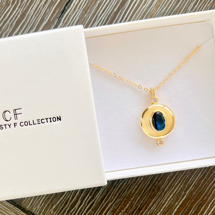 A gold pendant necklace with a dark blue oval stone and a tiny clear accent stone, displayed inside an open white jewelry box next to the branded lid. The box lid reads 'CRYSTAL F COLLECTION'.