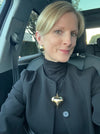 Woman sitting in a car wearing a dark coat with a heart-shaped pendant.