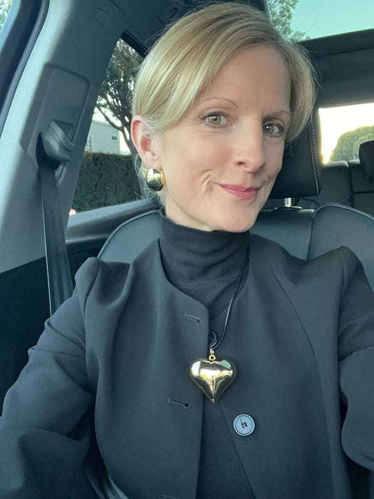 Woman sitting in a car wearing a dark coat with a heart-shaped pendant.